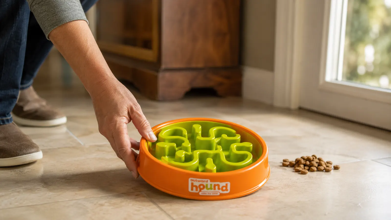 Hand placing the Outward Hound Fun Feeder Slo Bowl on a tile floor next to a measuring cup of kibble