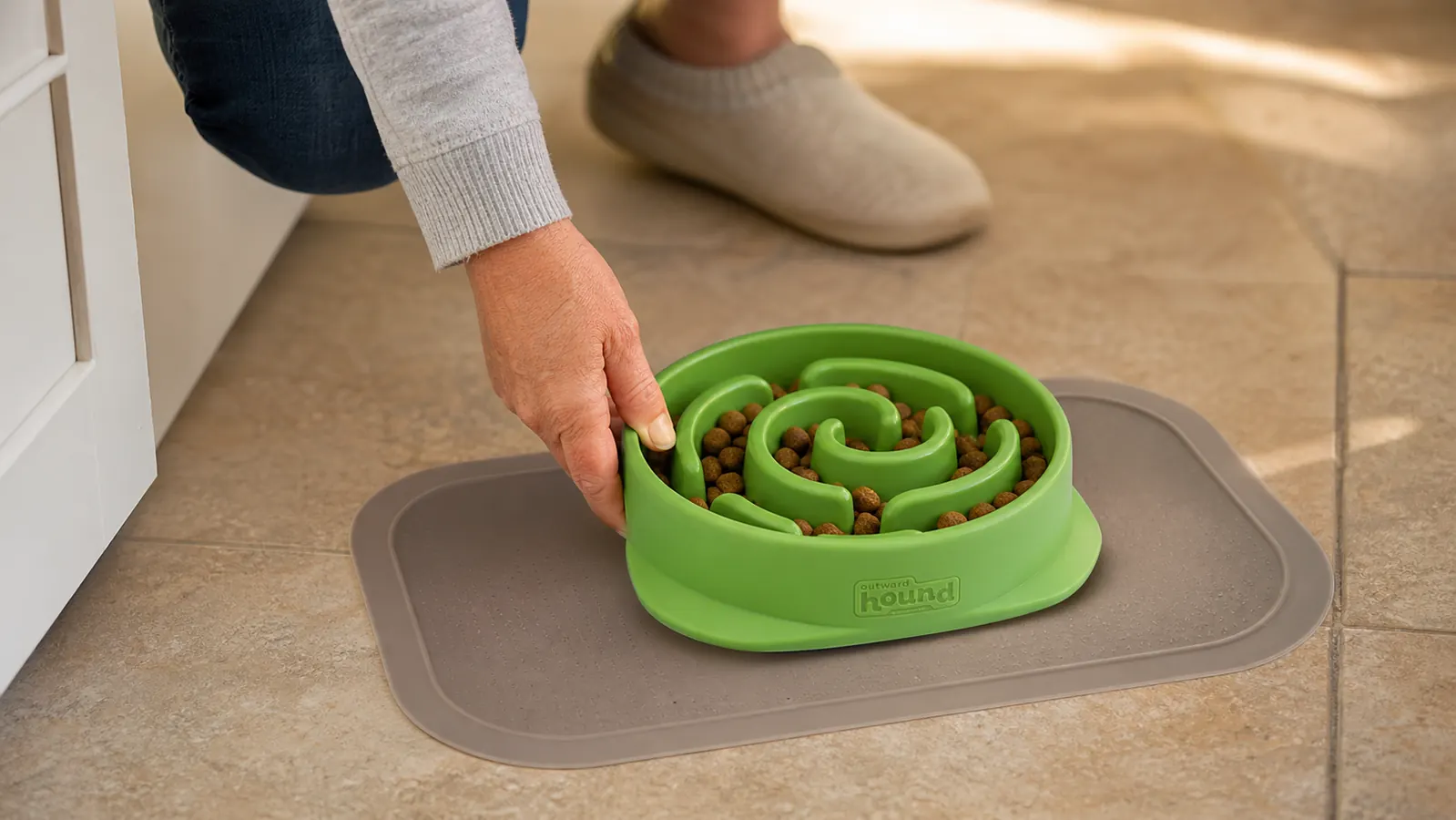Hand placing the Outward Hound Fun Feeder slow bowl on a floor mat before mealtime